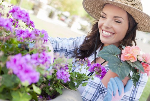A woman in a checkered shirt and wide-brimmed straw hat is tending to a garden filled with vibrant flowering plants, including purple, pink, and peach blossoms. She is using her gloved hands to carefully handle the flowers, which are situated in a patio or garden bed with lush green foliage and a backdrop of blurred outdoor scenery. The garden area appears well-maintained, with a mix of flowering perennials and greenery, and sunlight illuminating the scene. This outdoor space showcases a neatly organized garden typical of residential landscaping services offered in Waterloo, with a focus on planting and garden care. The image emphasizes a cheerful and healthy garden environment, aligned with professional gardening and lawn care services provided by Lawn Mowing Waterloo.